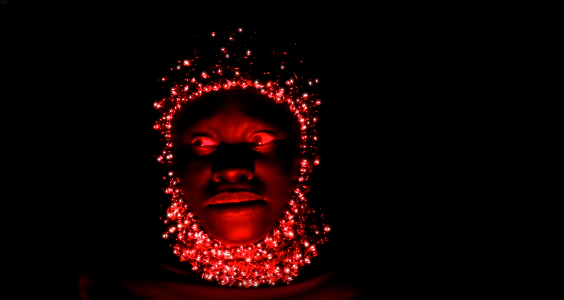 A portrait created by Shertise Solano. It is a black-and-red photograph of the face of a Black person. A balaclava made of metal bells covers the head. Red light shines on the face.
